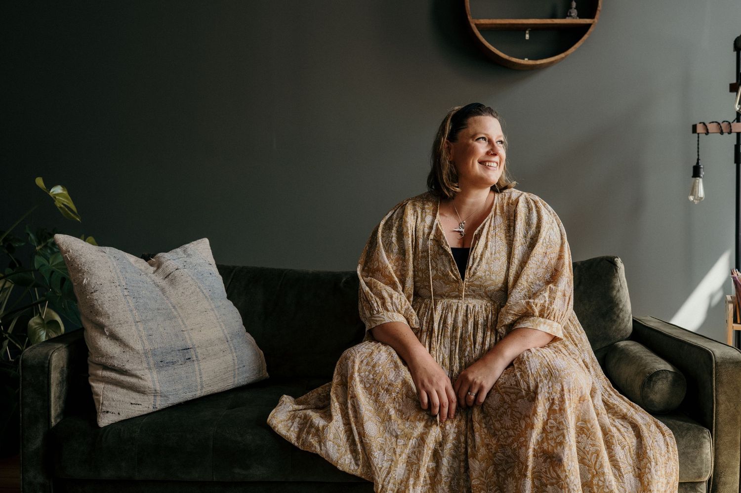 Woman in floral maxi dress sitting on green velvet sofa, smiling contentedly in a modern dark-walled living room.