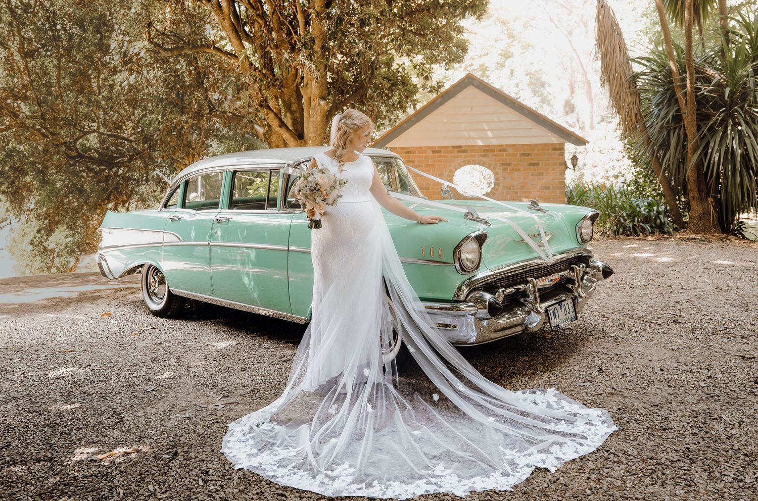 A bride in a flowing white dress poses beside a mint green vintage car during a rustic outdoor photoshoot.