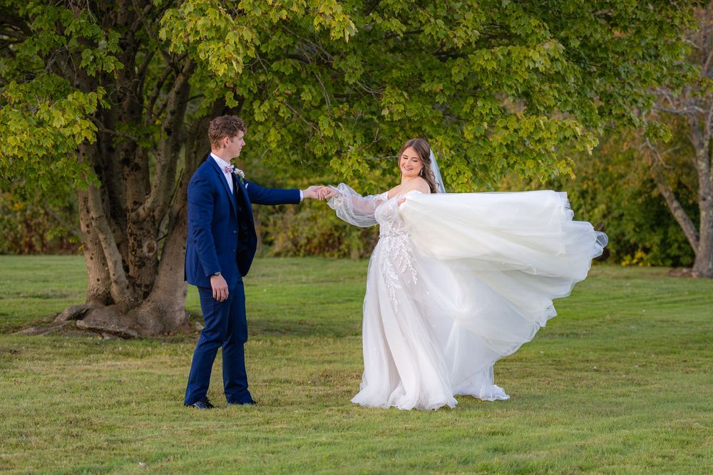 A couple in wedding attire dances on a green lawn with autumn trees in the background.