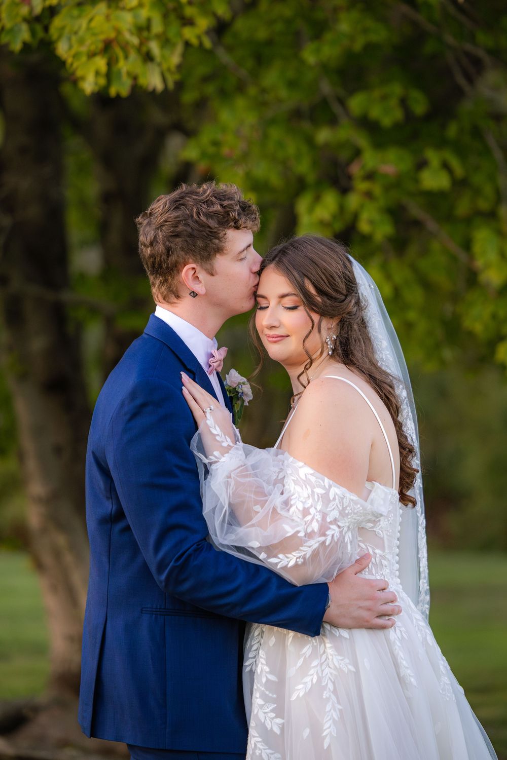 A romantic wedding moment between couple in navy suit and white lace dress amid lush green garden setting.