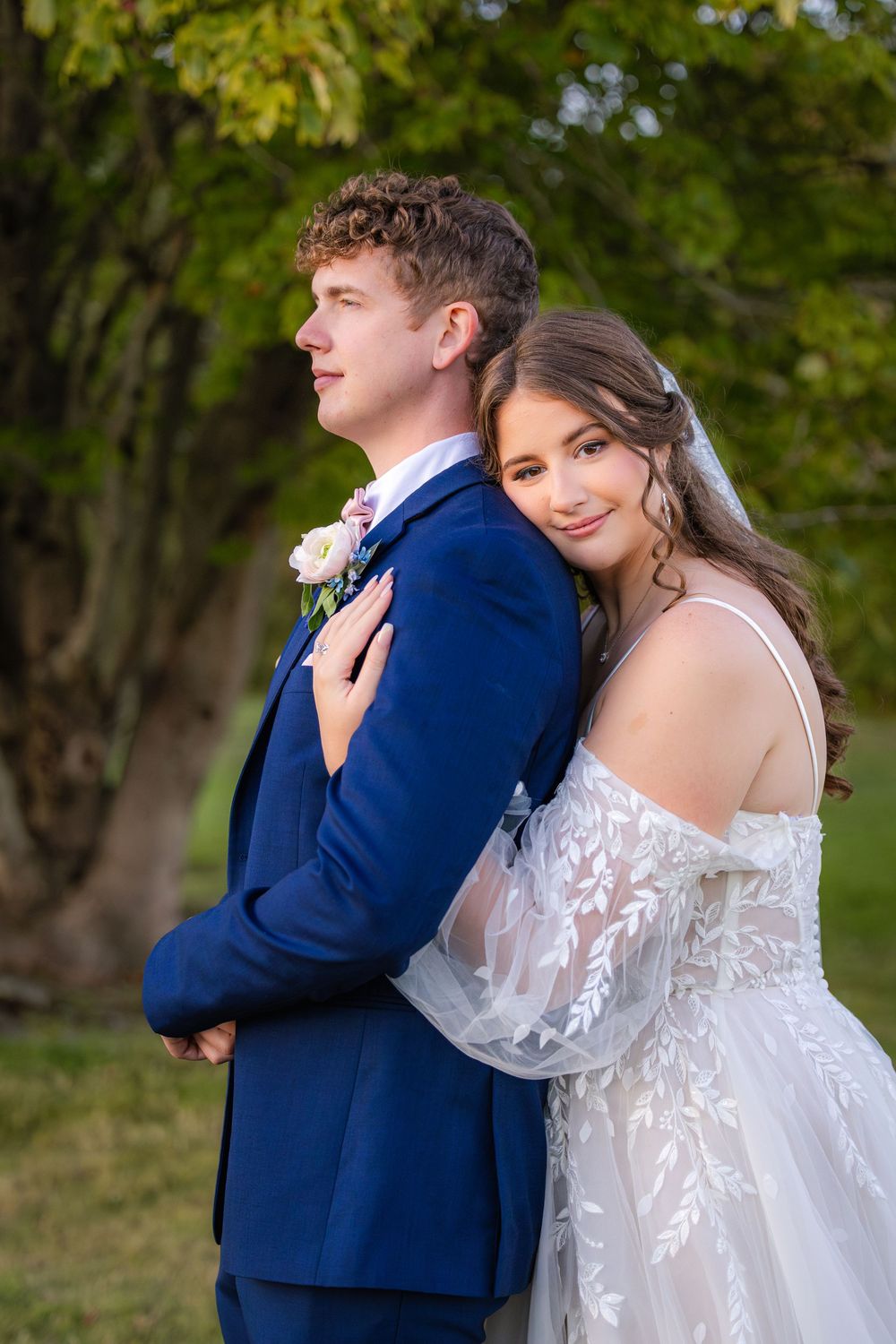 A couple in wedding attire, with a bride in lace gown and groom in navy suit, embracing outdoors among autumn trees.