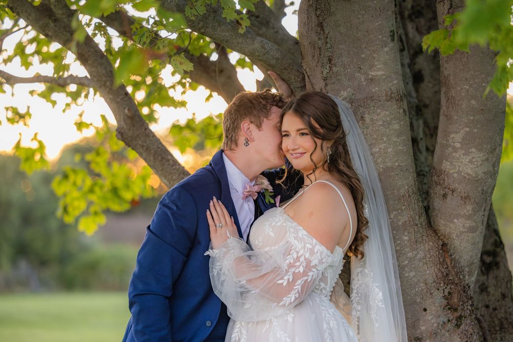 A bride and groom share a romantic kiss under a tree's canopy during their outdoor wedding photoshoot.