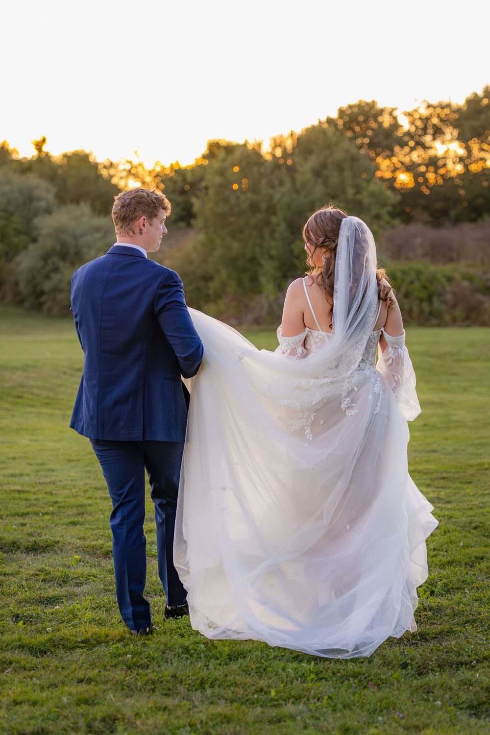 A wedding couple walks through a grassy field during golden hour while the bride's elegant white veil flows in the breeze.
