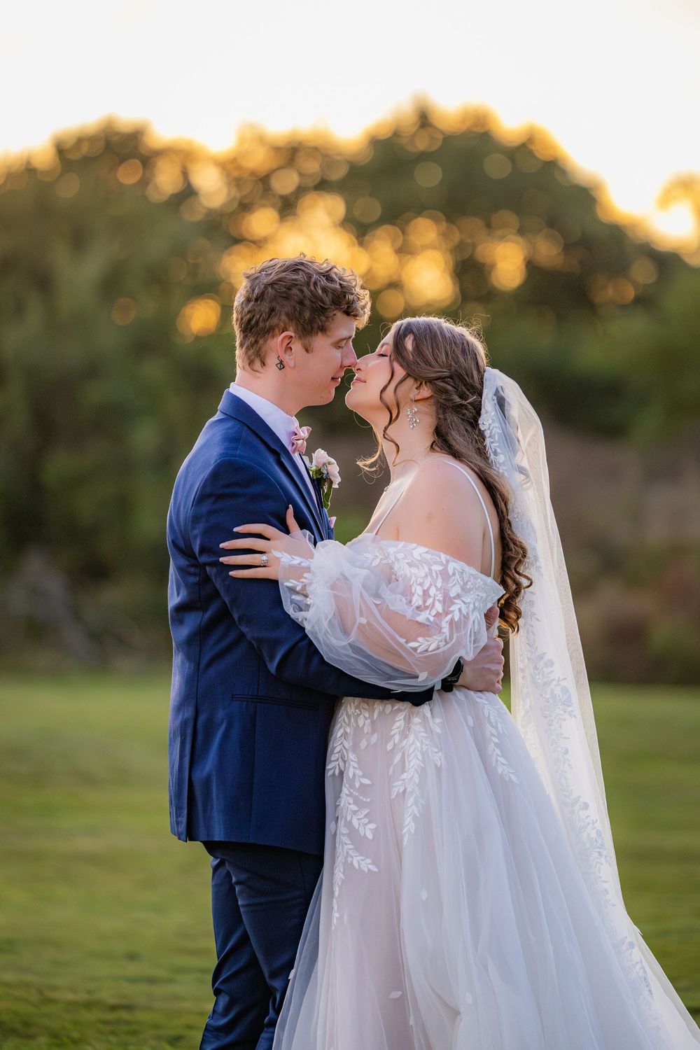 A romantic wedding couple embraces outdoors during golden hour while wearing a navy suit and lace gown.