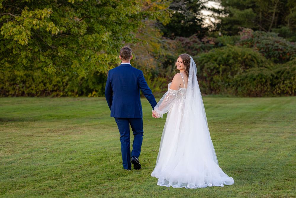 A couple walks across a sun-dappled lawn wearing formal wedding attire in a serene outdoor setting.