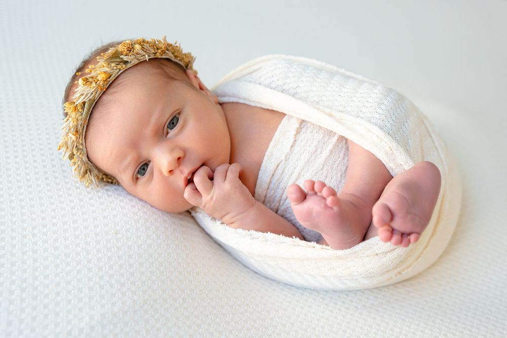 A newborn baby swaddled in white fabric lying on a light background while wearing a gold crown headband.