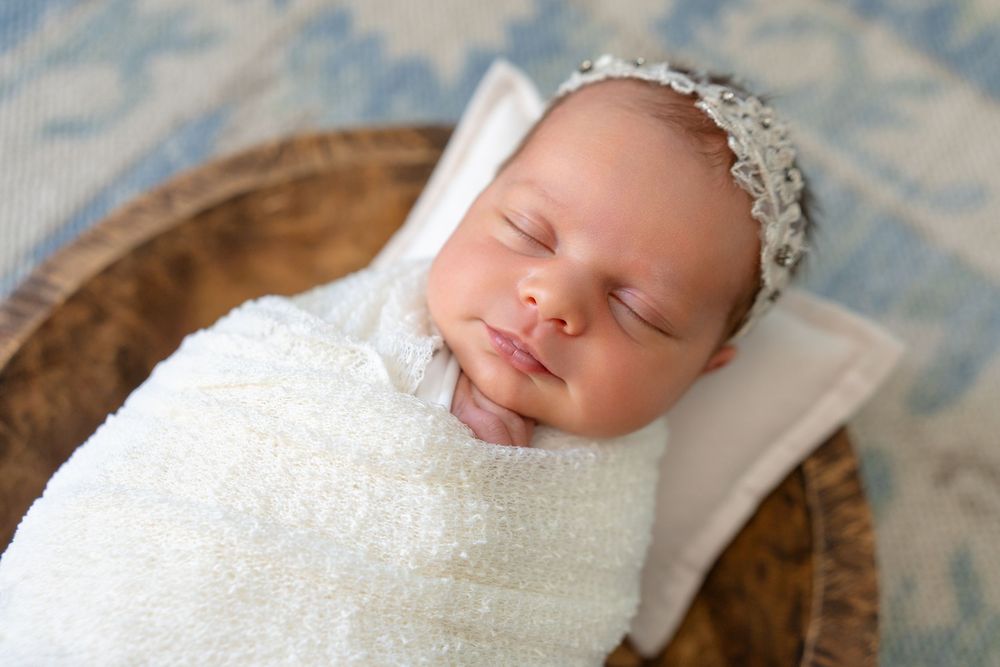 A sleeping newborn wearing a white lace bonnet and swaddle with arms tucked in rests peacefully against a textured backdrop.