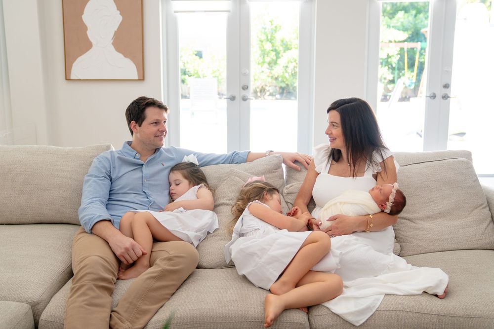 Parents cuddling with newborn baby on couch in bright living room.