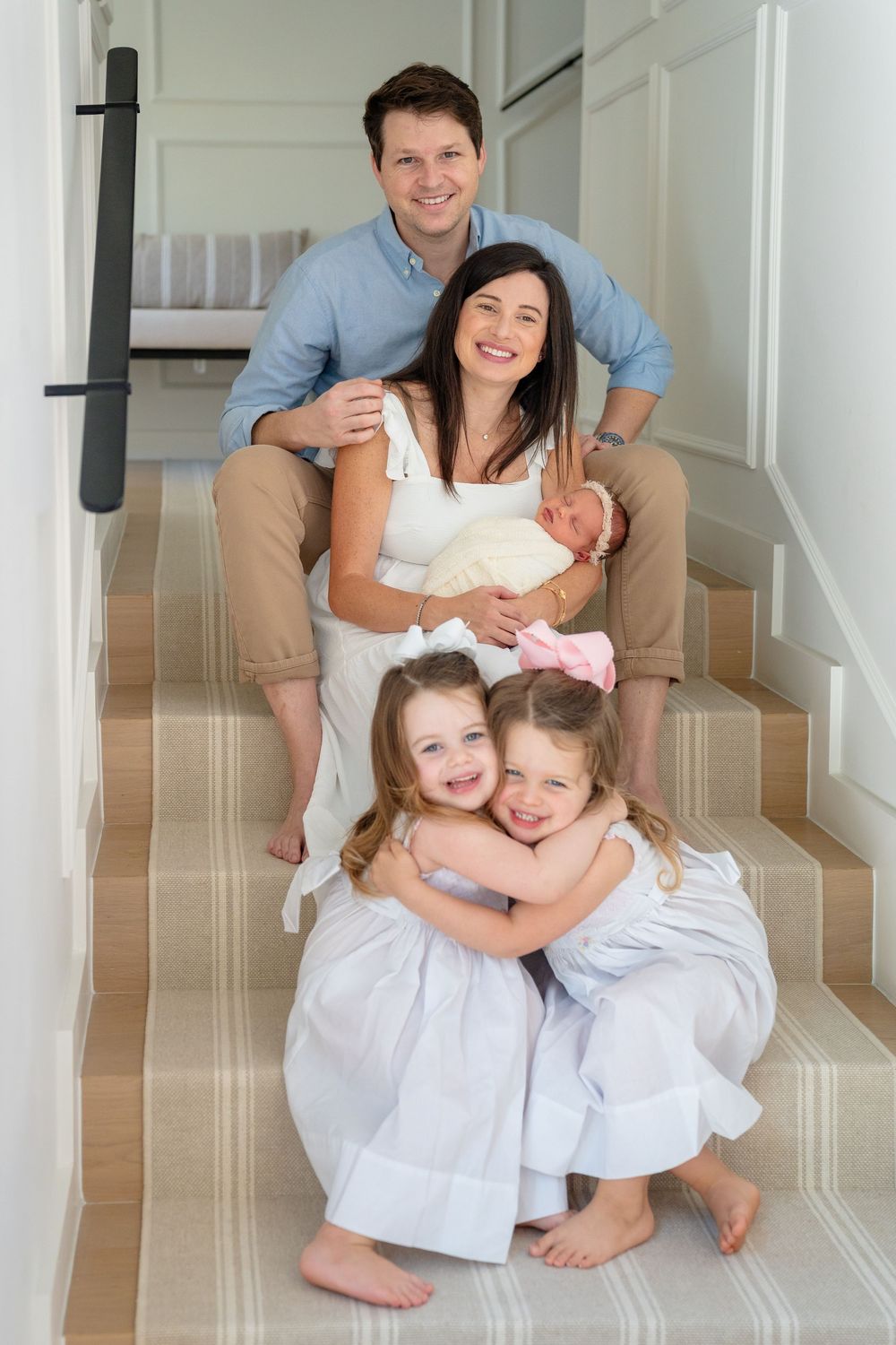 Family of five in coordinating white outfits share joyful embraces while seated on carpeted stairs.