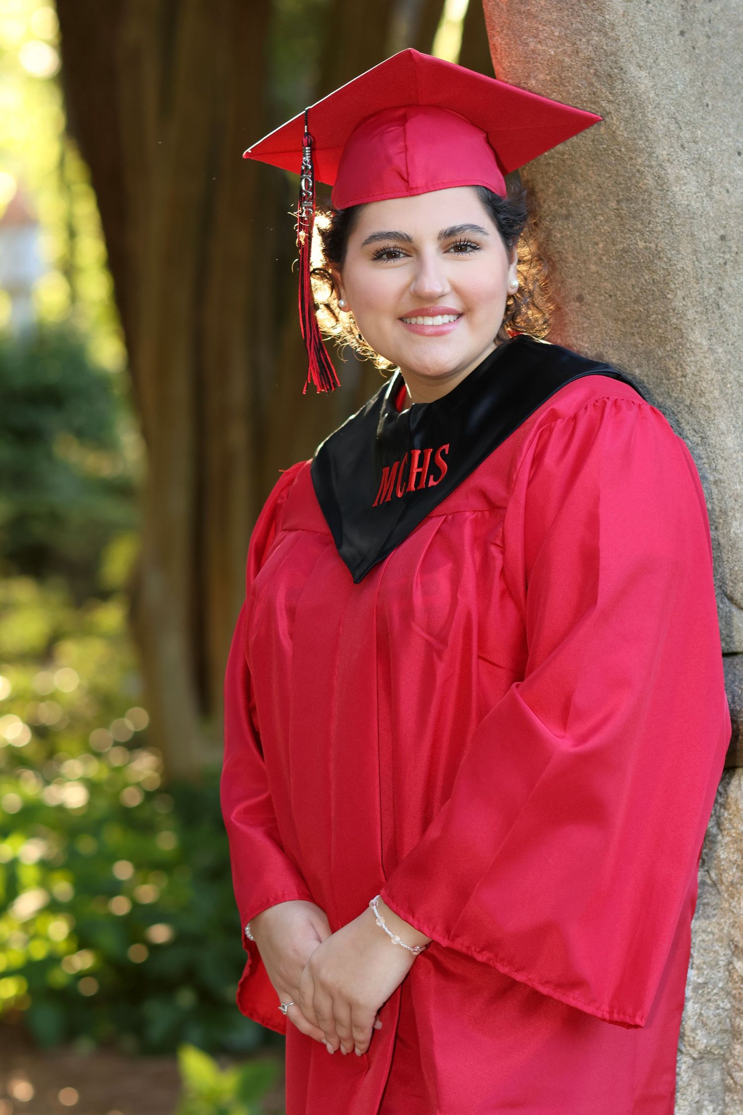High school senior girl leaning against a rock at sunset, the light illuminating her hair from behind wearing a red graduation robe.