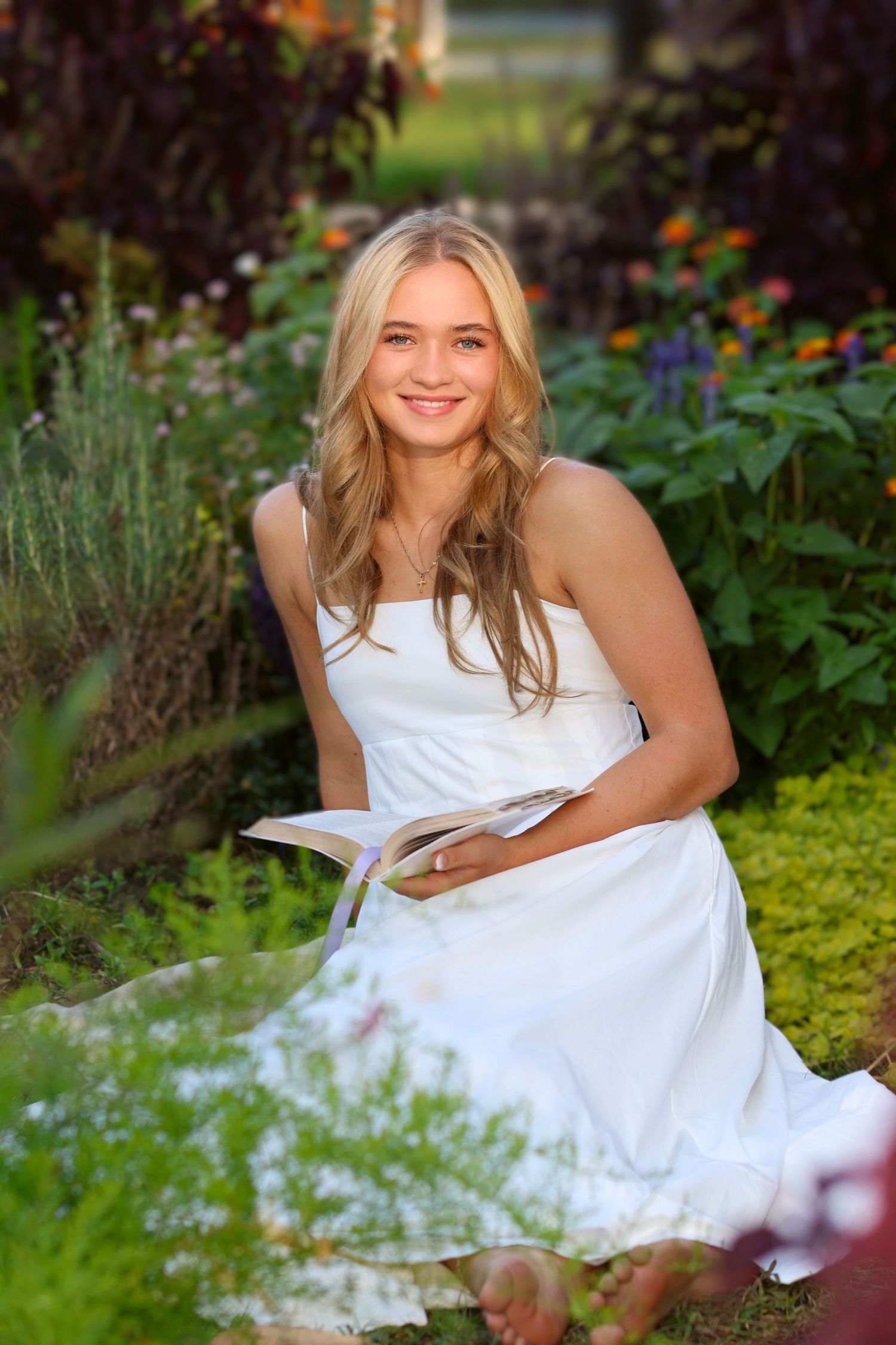 High school senior girl in a casual white dress surrounded by beautiful flowers holding a Bible, the setting sunlight illuminating her face.