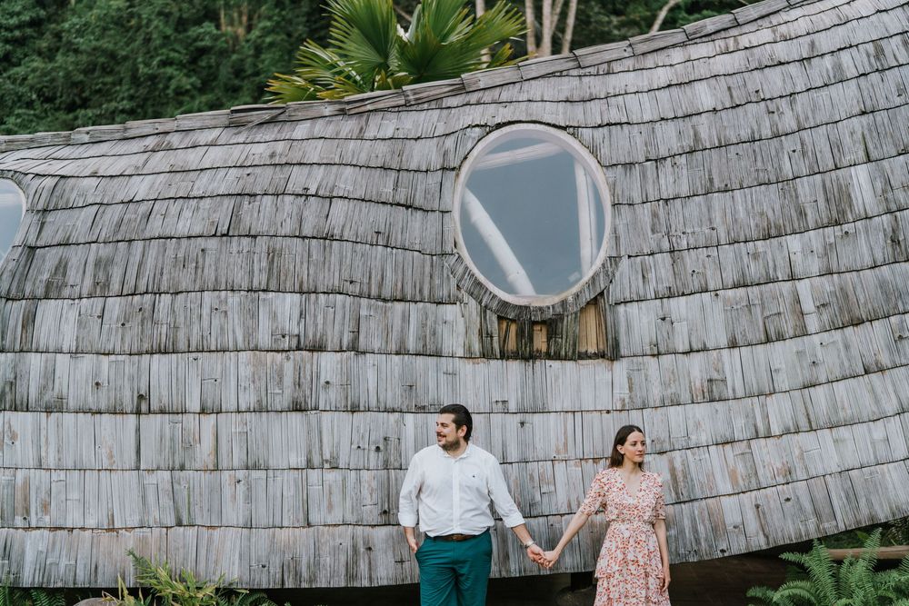 A couple holds hands in front of a curved bamboo wall with circular windows.