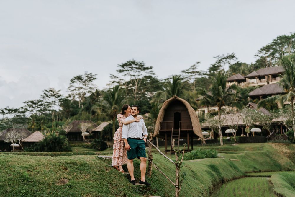 A couple stands on a green hillside with traditional bamboo architecture in the distance.