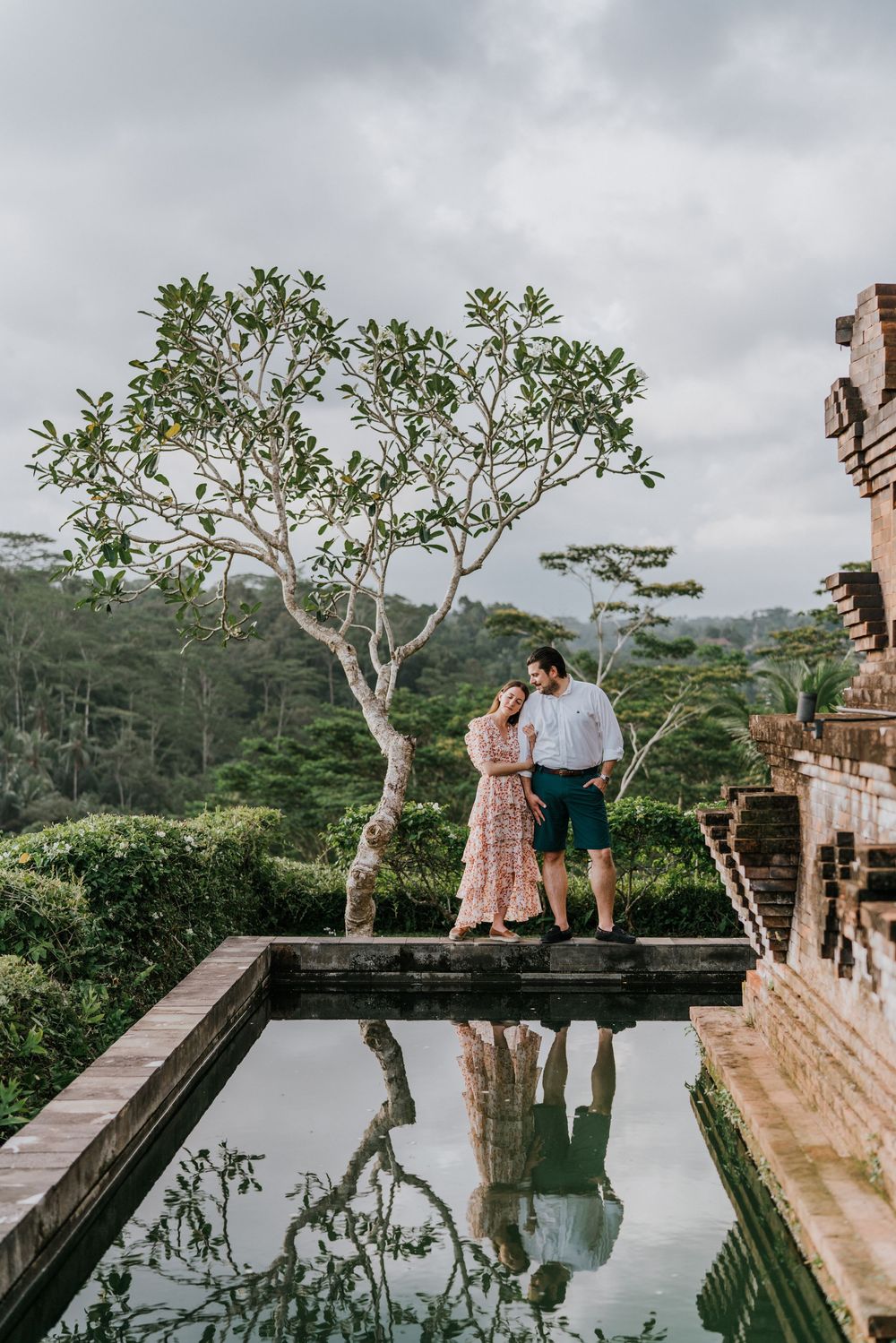 A couple shares an intimate moment by a reflective pool with tropical trees in the background.