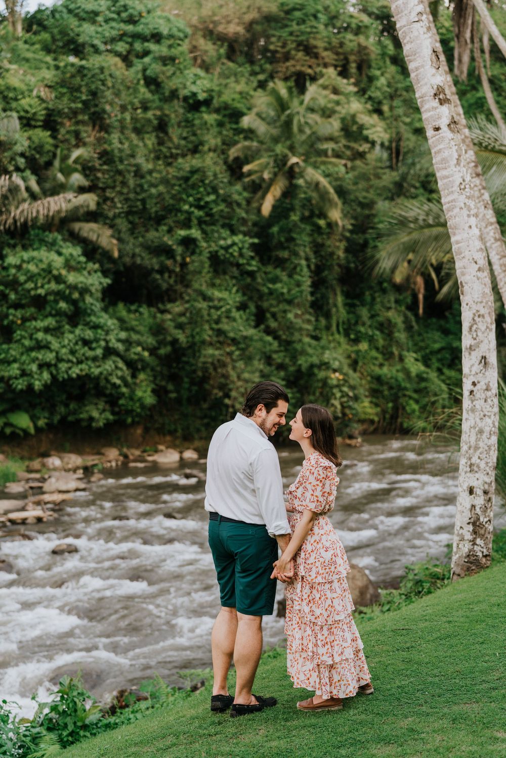A tender moment captured beside a rushing river with palm trees in the background.