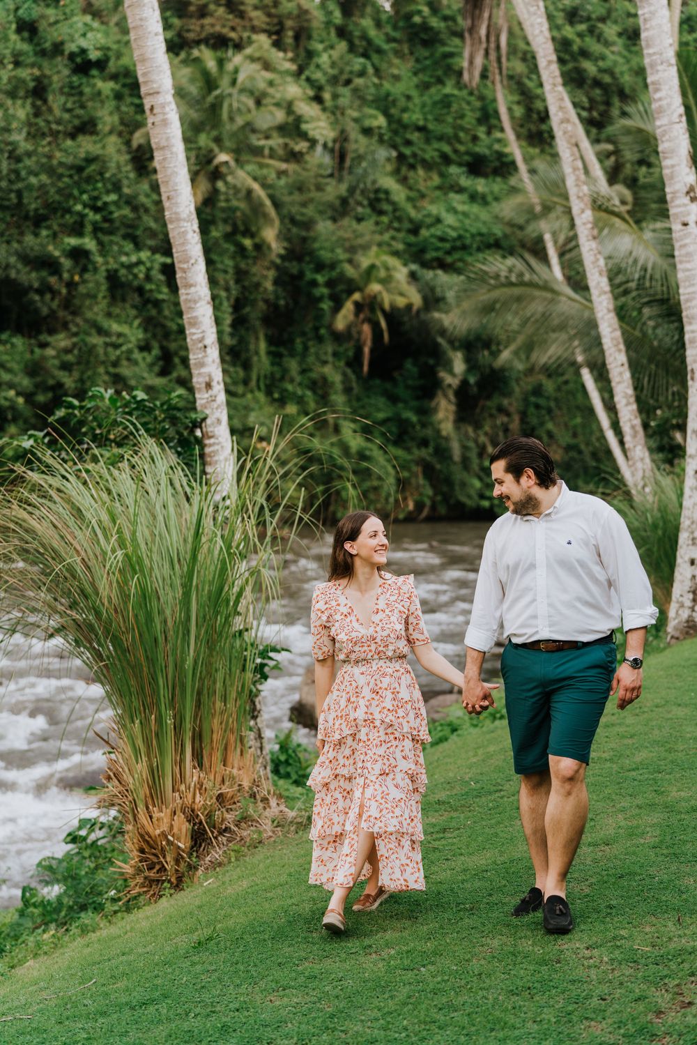 A couple walks along a grassy riverbank lined with palm trees and tropical plants.
