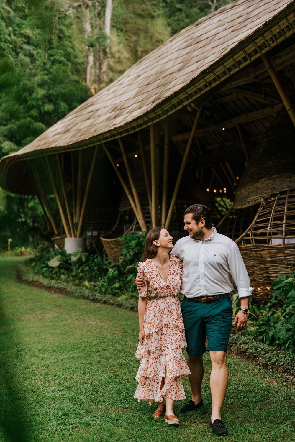 A couple stands together by a rustic bamboo building with a thatched roof in a tropical setting.