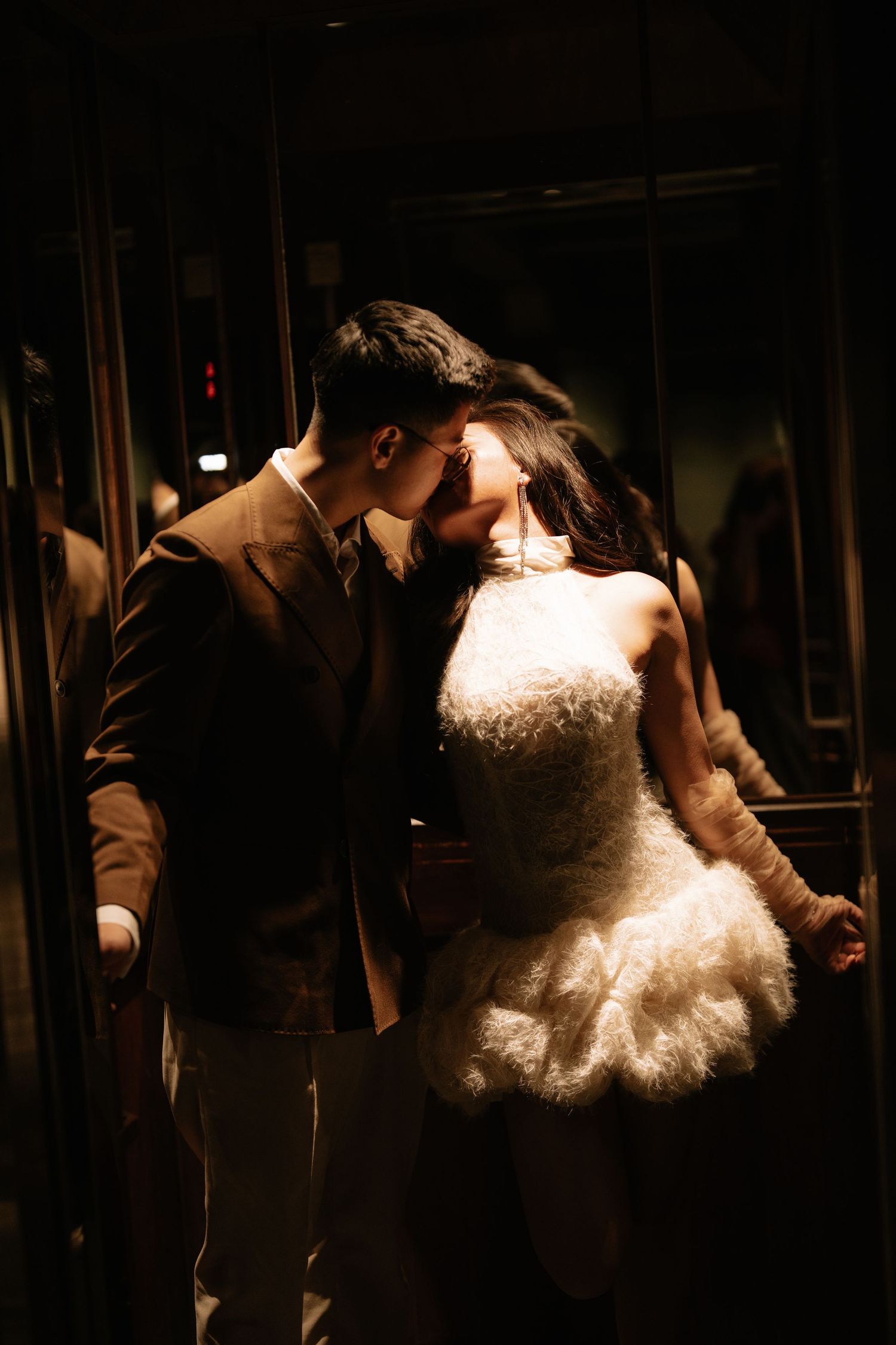 Bride and groom share a romantic kiss in a dimly lit elevator, bride wearing a fluffy white mini wedding dress.