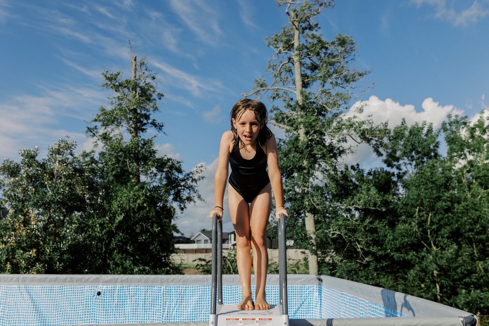 A swimming pool with diving board against blue summer sky surrounded by green trees.