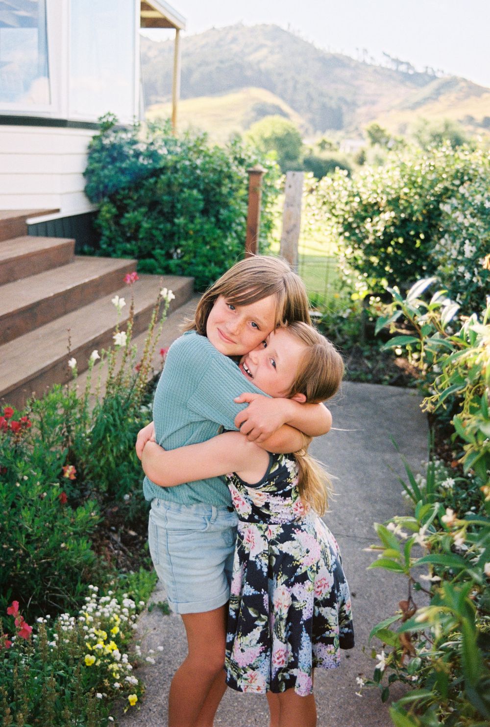 Two children sharing a warm hug among blooming flowers in a garden with mountains in the background.