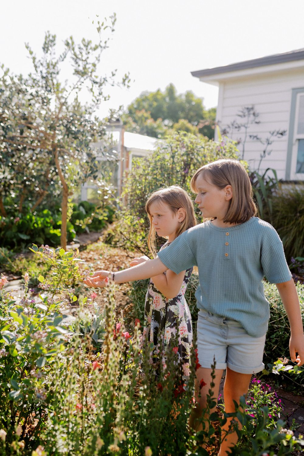 Kids exploring a backyard garden filled with blooming flowers near a white house.