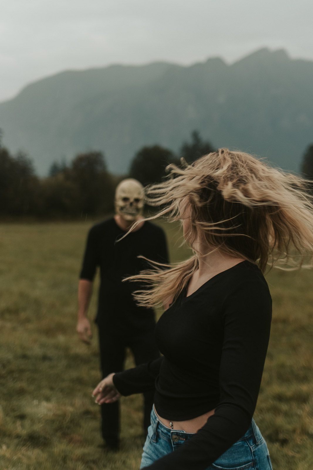 Spooky Halloween couple photoshoot with woman in black top, jeans turning away as masked guy following in foggy field, spokane photographer