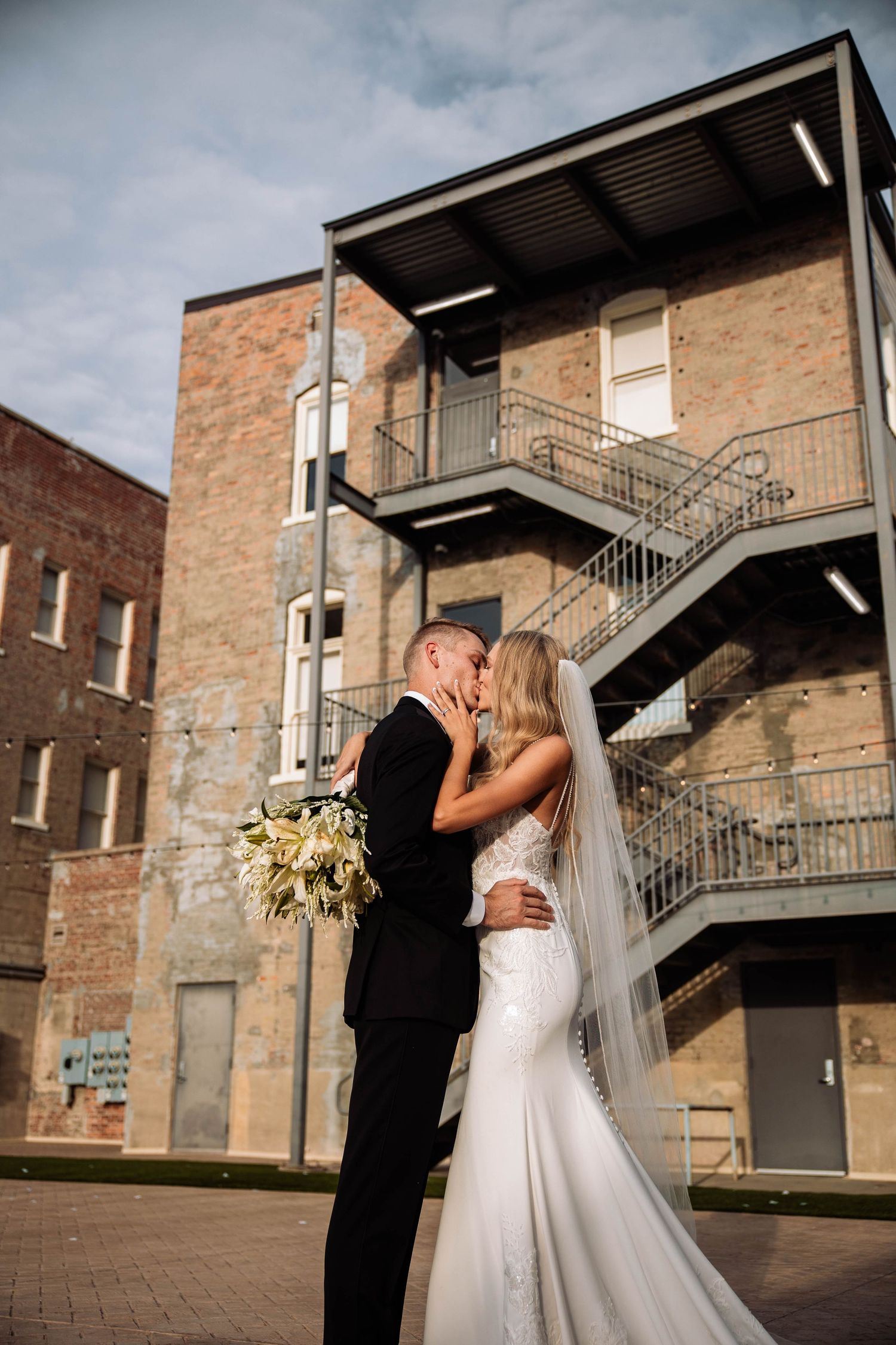 Wedding couple embraces against urban brick backdrop with metal fire escape.