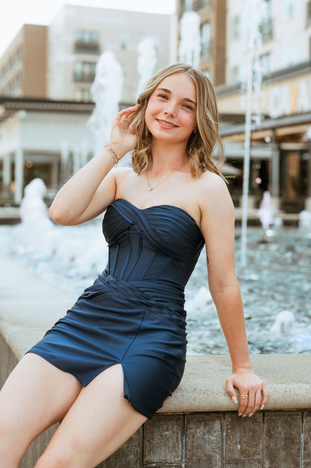 Woman in navy blue swimsuit poses elegantly by fountain in a warm outdoor setting.