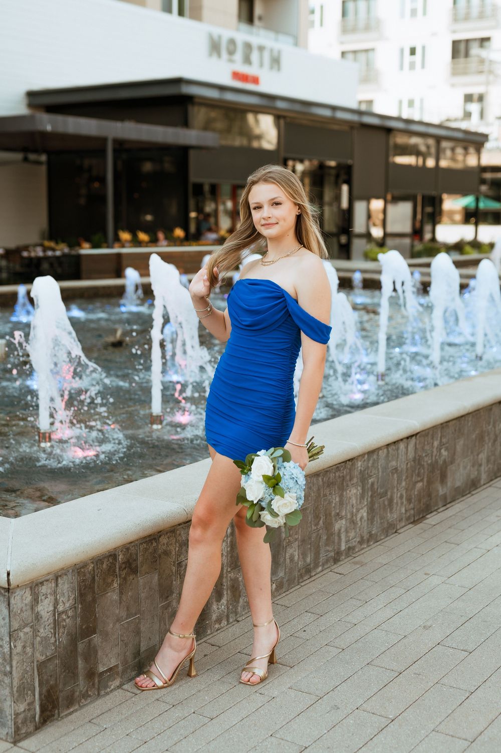 Woman wearing blue dress and gold sandals stands near modern fountain with white flowers in hand.
