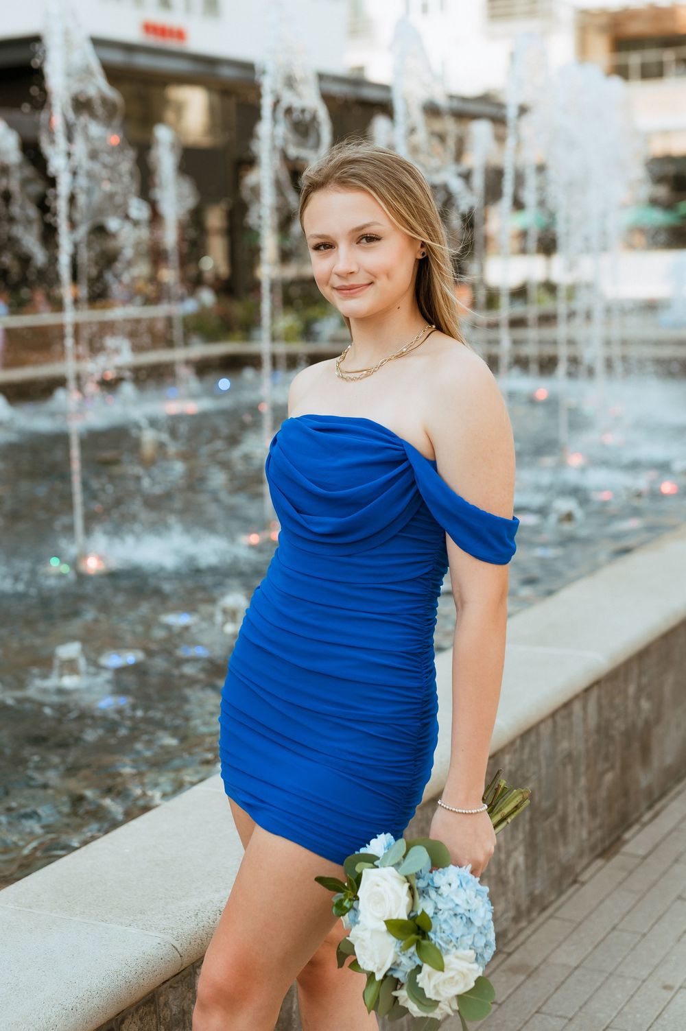 A woman in a royal blue one-shoulder dress poses by a fountain holding light blue and white flowers.