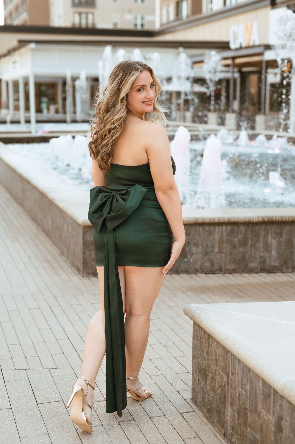 Woman in an elegant dark green dress with bow detail poses against urban backdrop in high heels.