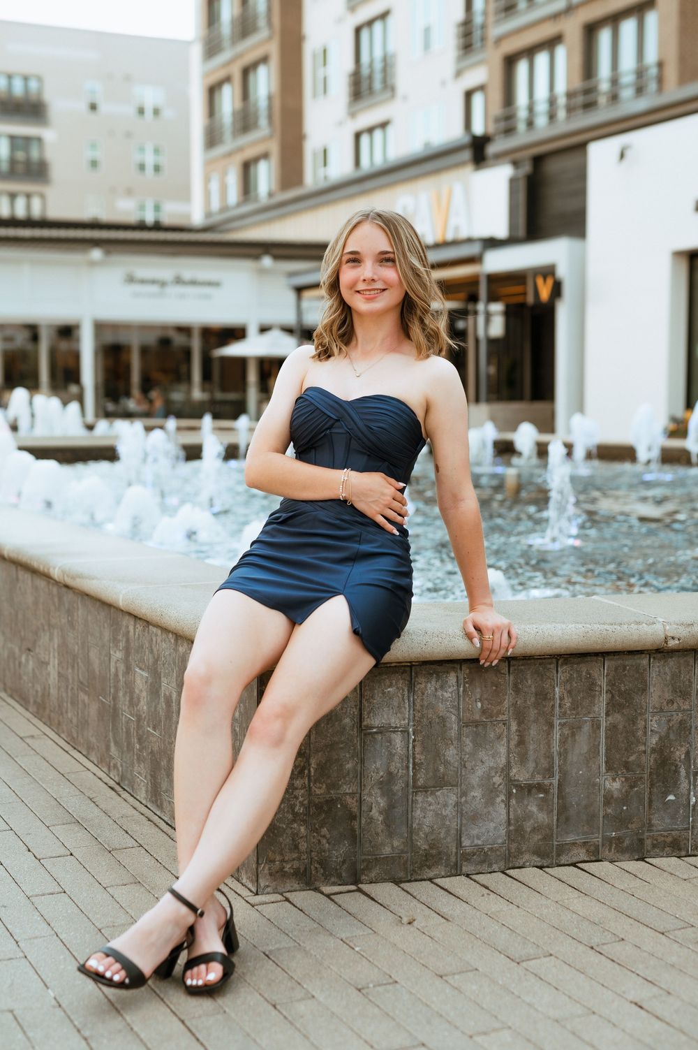 A woman in a navy blue dress sits casually on a stone ledge near fountains in an urban setting.