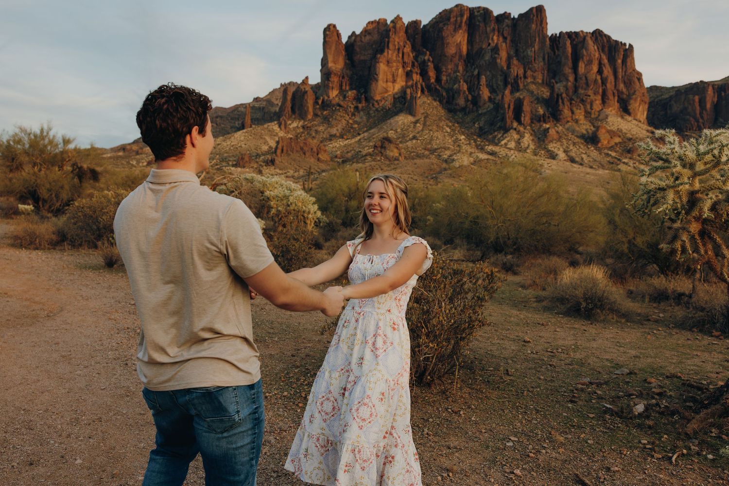A couple stands on a desert trail with the majestic Superstition Mountains rising in the background at sunset.