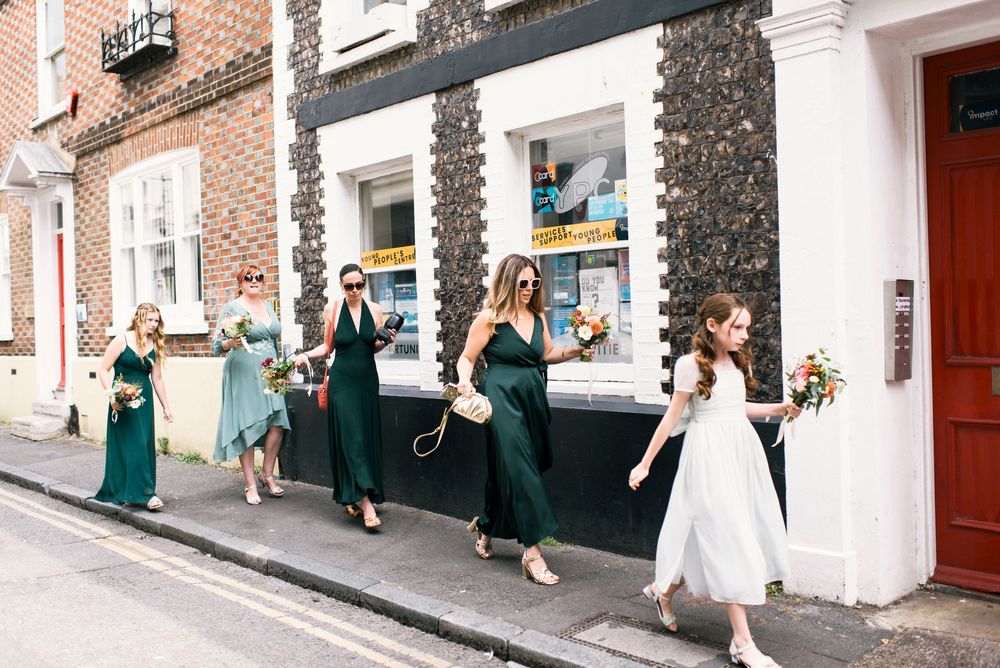 Bridesmaids in emerald green dresses walk down a sunny street in Brighton alongside a bride in white holding a colorful bouquet.