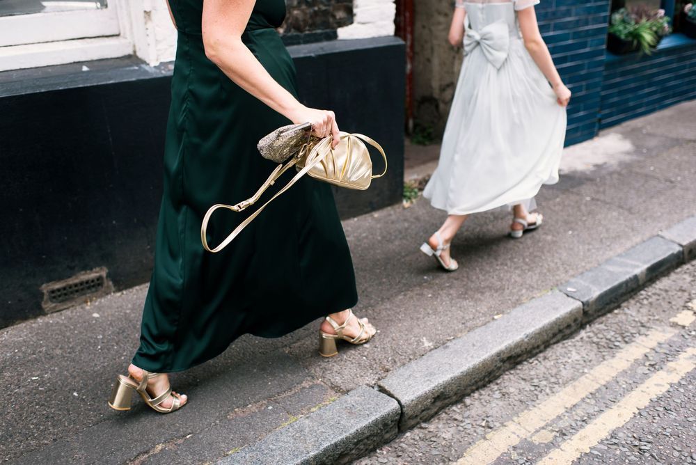 Two women in elegant dresses and gold heels walking on a sidewalk, one holding a gold clutch purse.