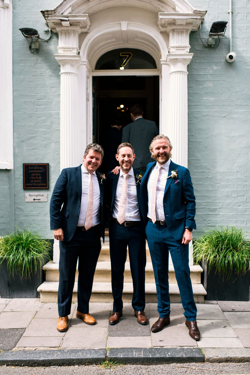 Three groomsmen in navy suits smiling outside elegant white doorway of the Hotel Du Vin Brighton.