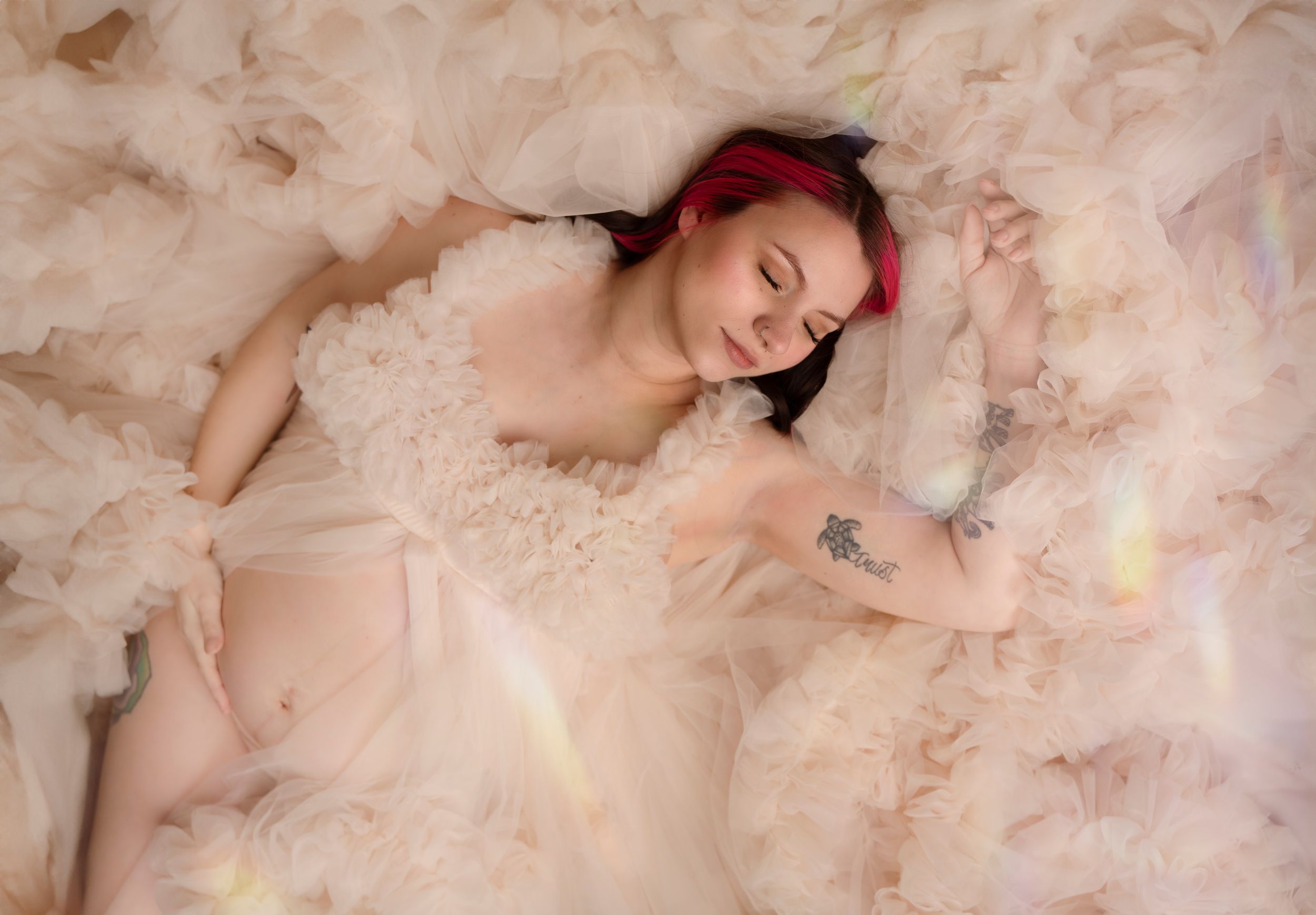 A woman in a flowy nude colored dress poses ethereally on beige fabric in a dreamy boudoir setting.