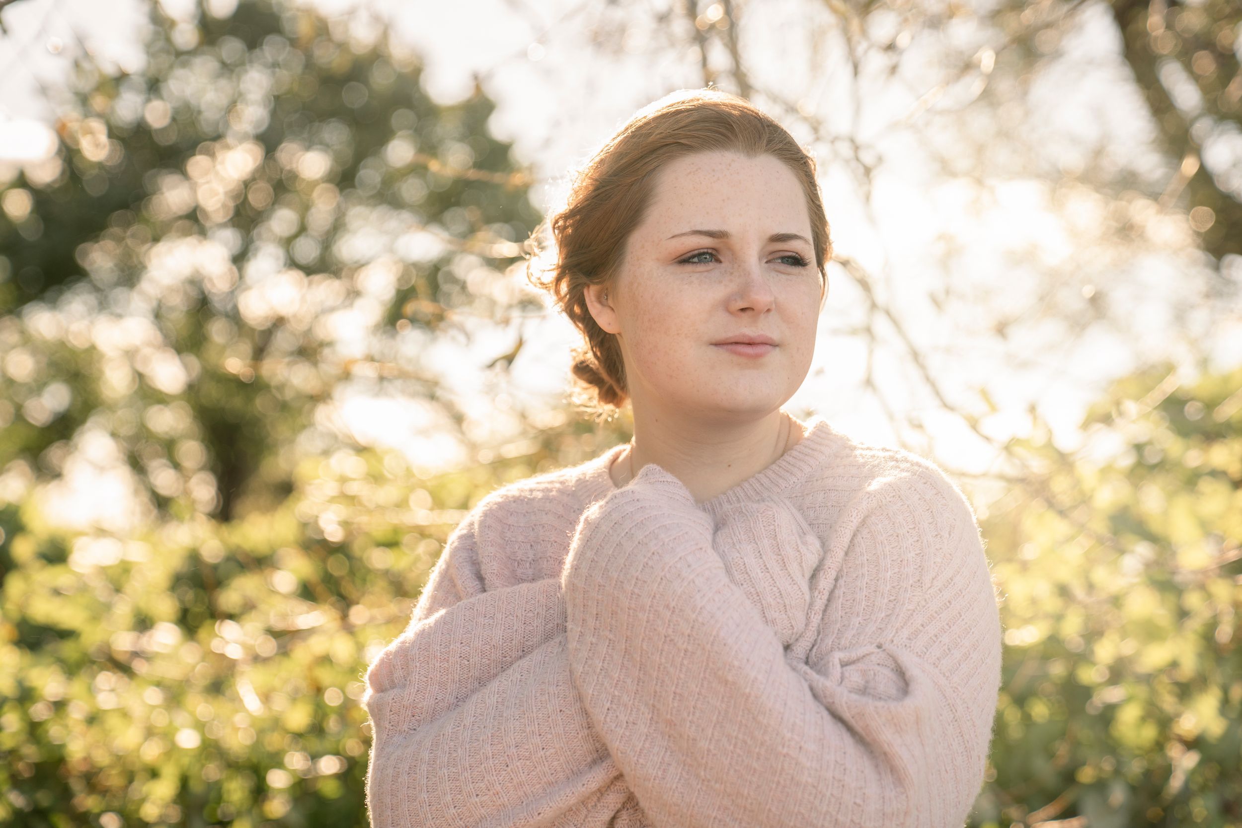 A person in a cozy sweater poses outdoors during golden hour sunlight with a soft natural backdrop.