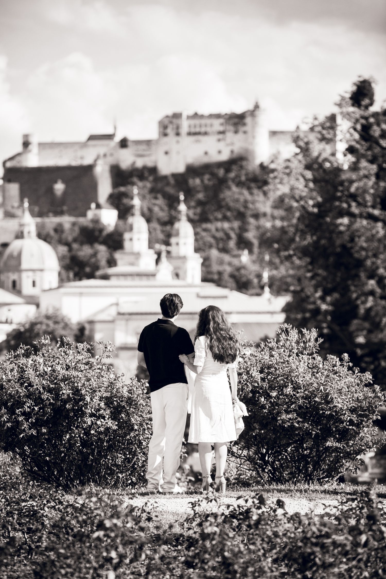 A black and white sequence showing two figures strolling among shrubbery with white structures visible in the background.