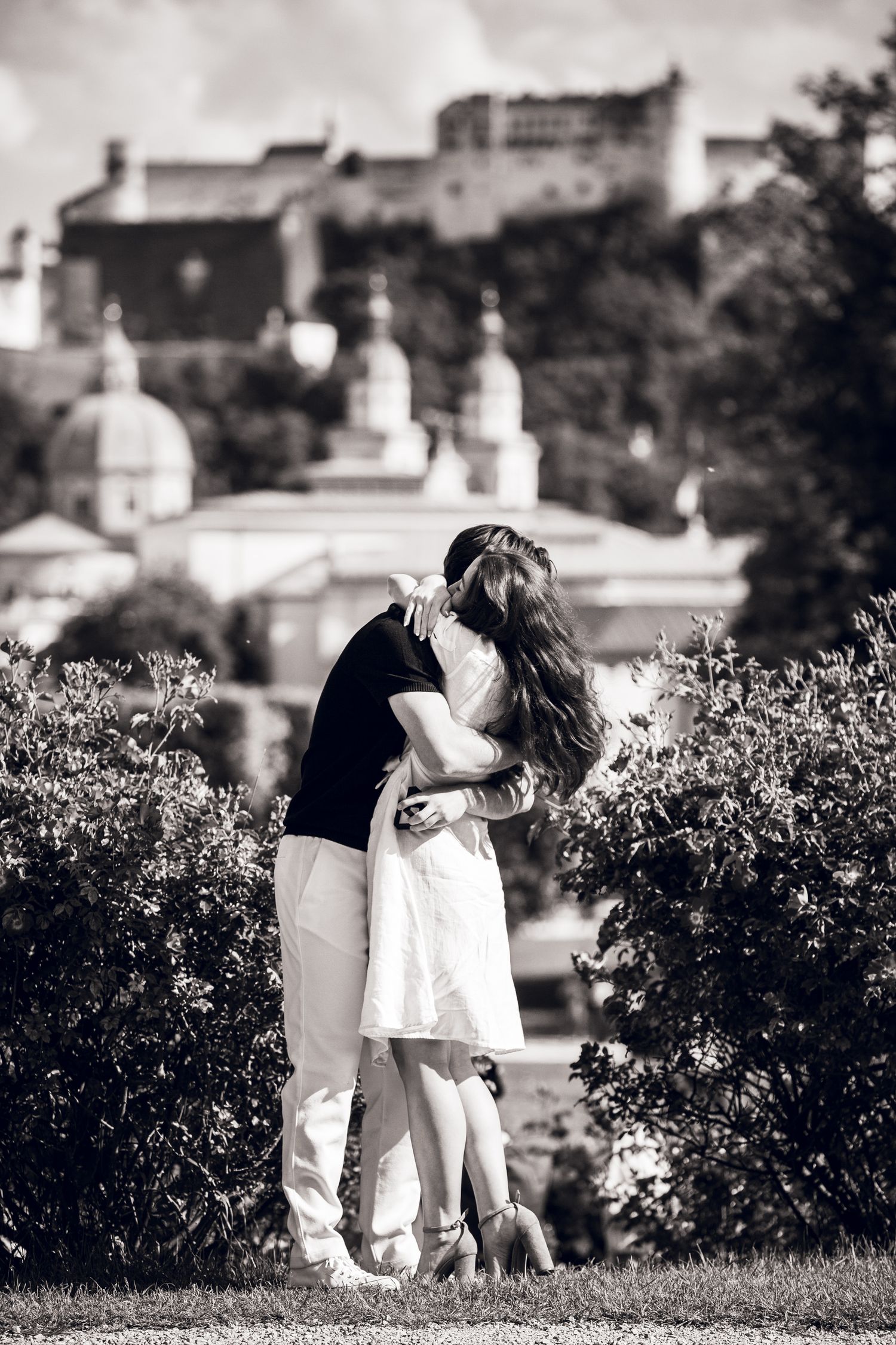 Black and white photo series showing a couple in casual clothing embracing outdoors near bushes on a sunny day.