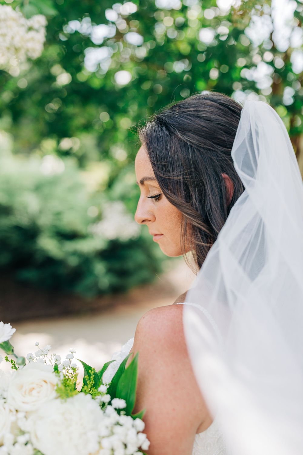 A profile view of a bride wearing a delicate white veil against a soft green natural backdrop with bokeh effects.