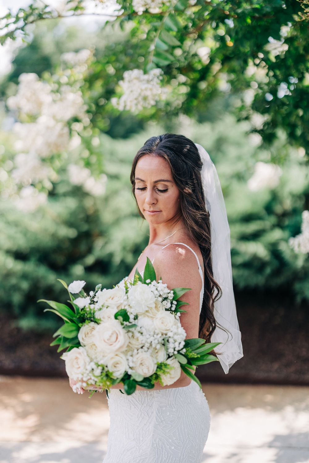 Bride in lace wedding dress holds elegant white floral bouquet against green garden backdrop.