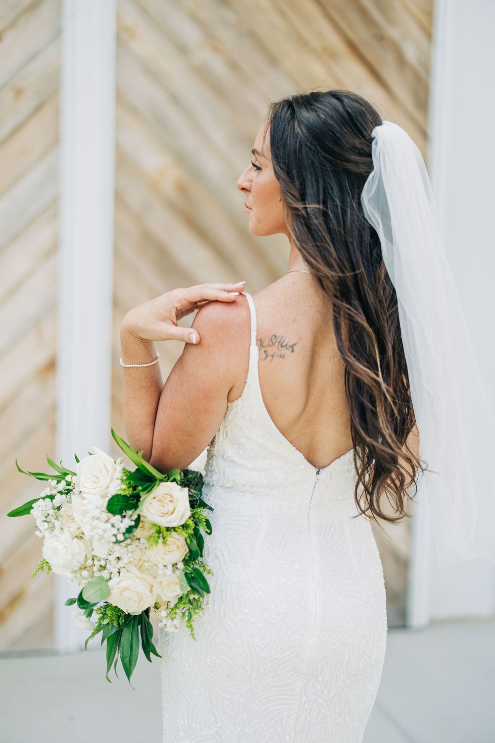 Bride in white wedding dress holds elegant bouquet of white roses and greenery against a cream striped wall.
