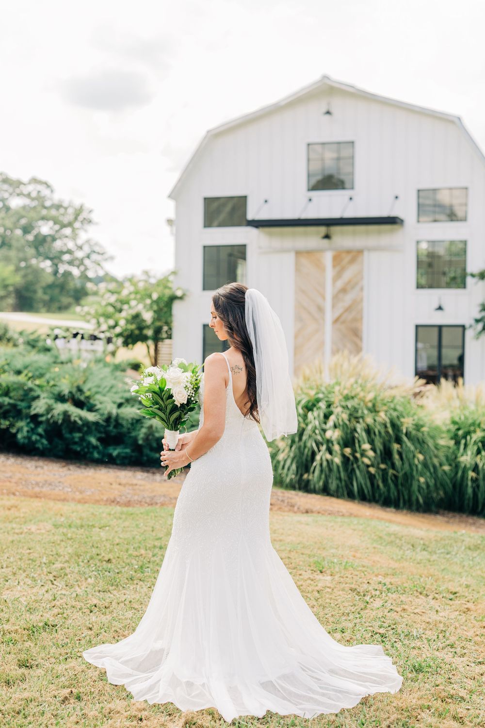 A bride in a flowing white wedding dress stands on a grassy hill with white barns in the background on a sunny day.