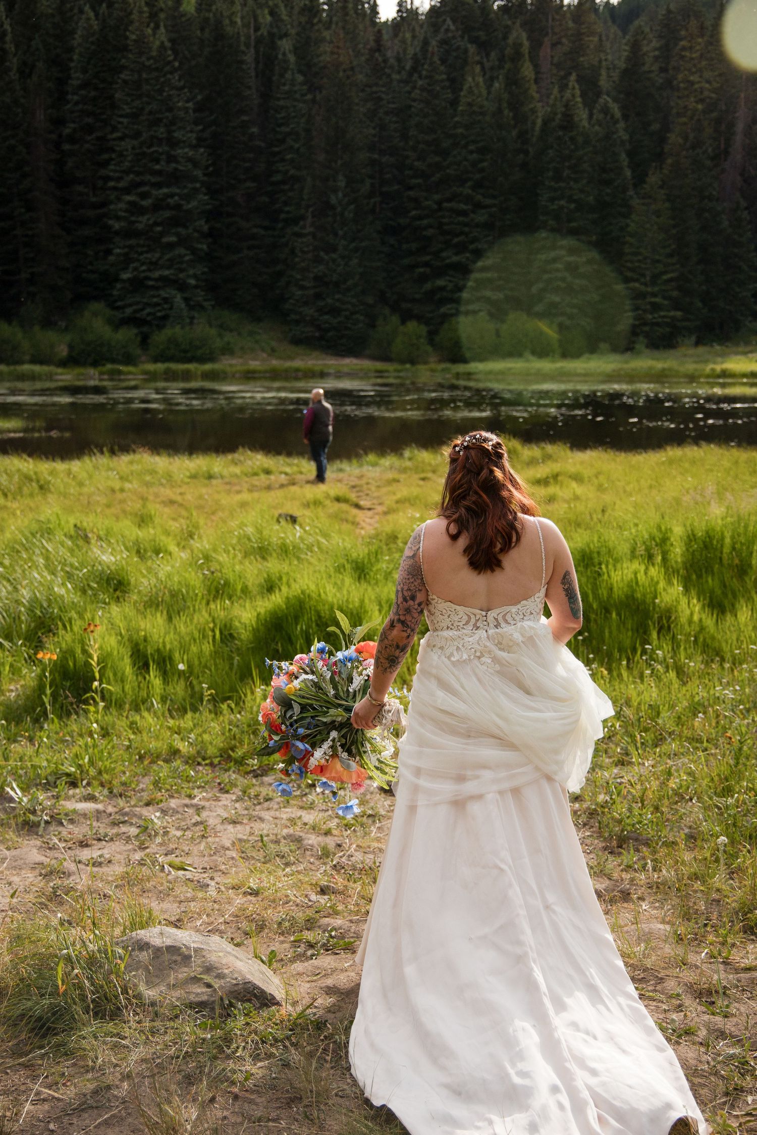A person in a white wedding dress walks through a scenic mountain meadow surrounded by evergreen forest.