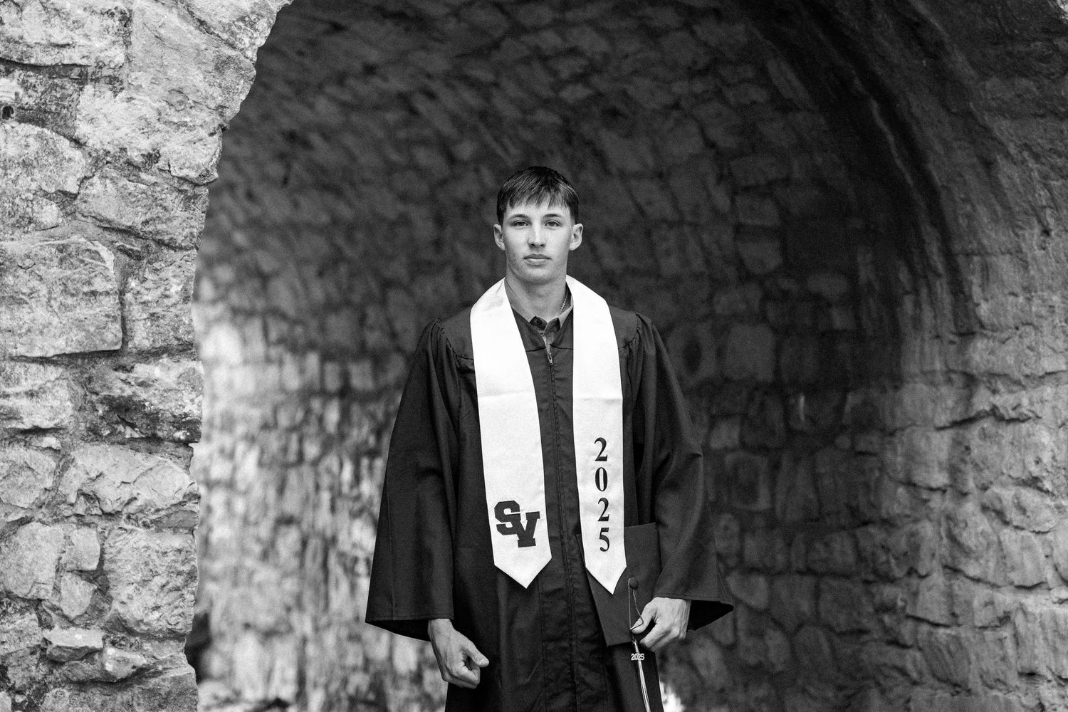 Graduate in black academic regalia and stole standing under stone archway in black and white photo.