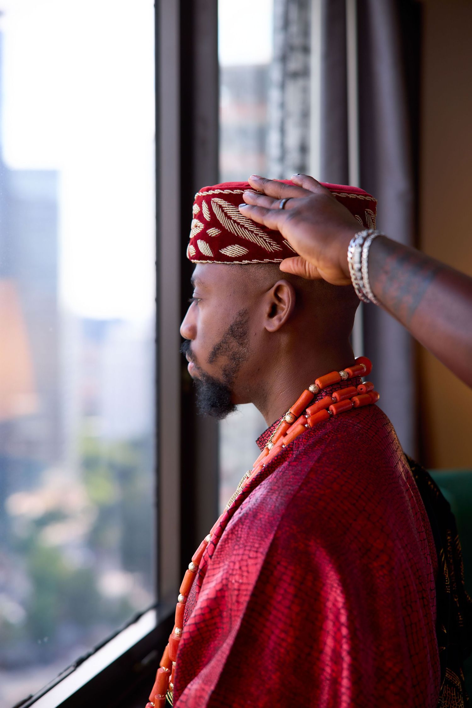 Someone adjusts traditional red and gold headwear while standing near large windows in an urban setting.