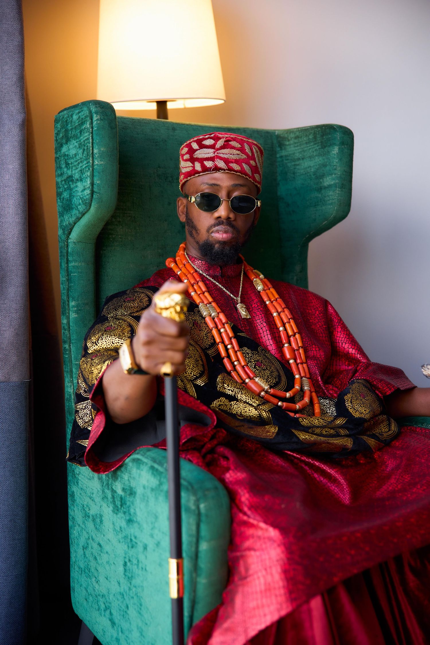 A figure in traditional Nigerian attire with red and gold robes sits regally in an emerald green velvet armchair beside a lamp.