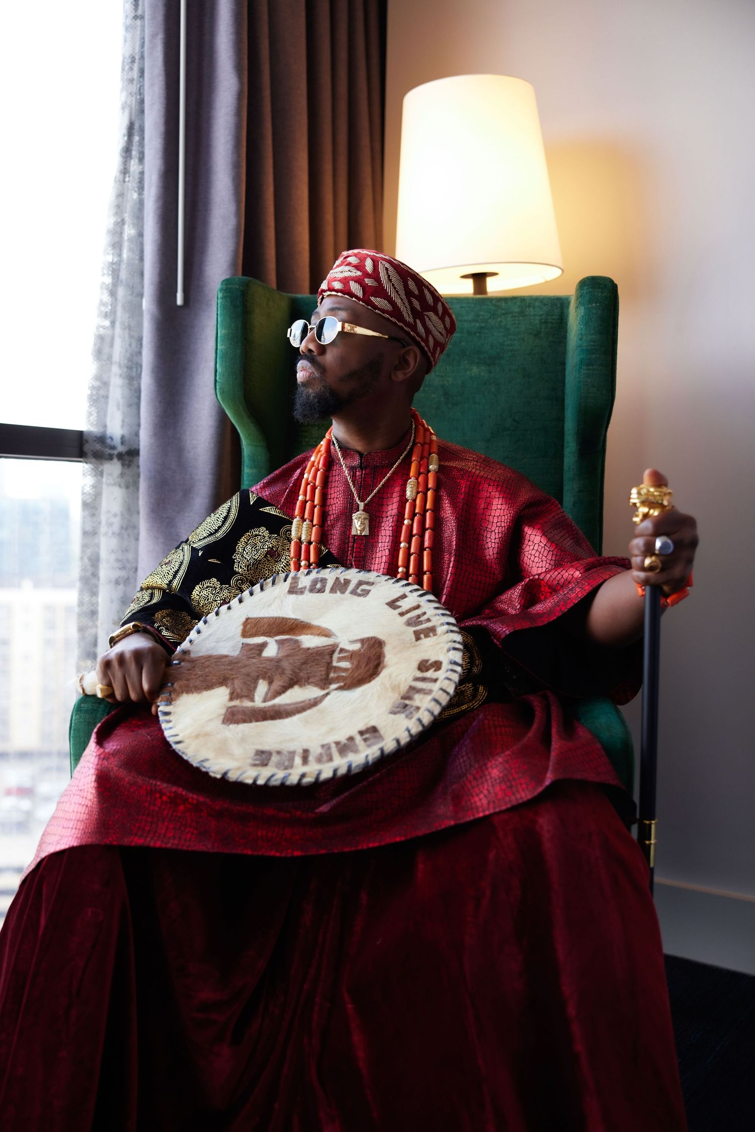 A musician in a red outfit plays a banjo while seated in a green armchair near a window with natural lighting.