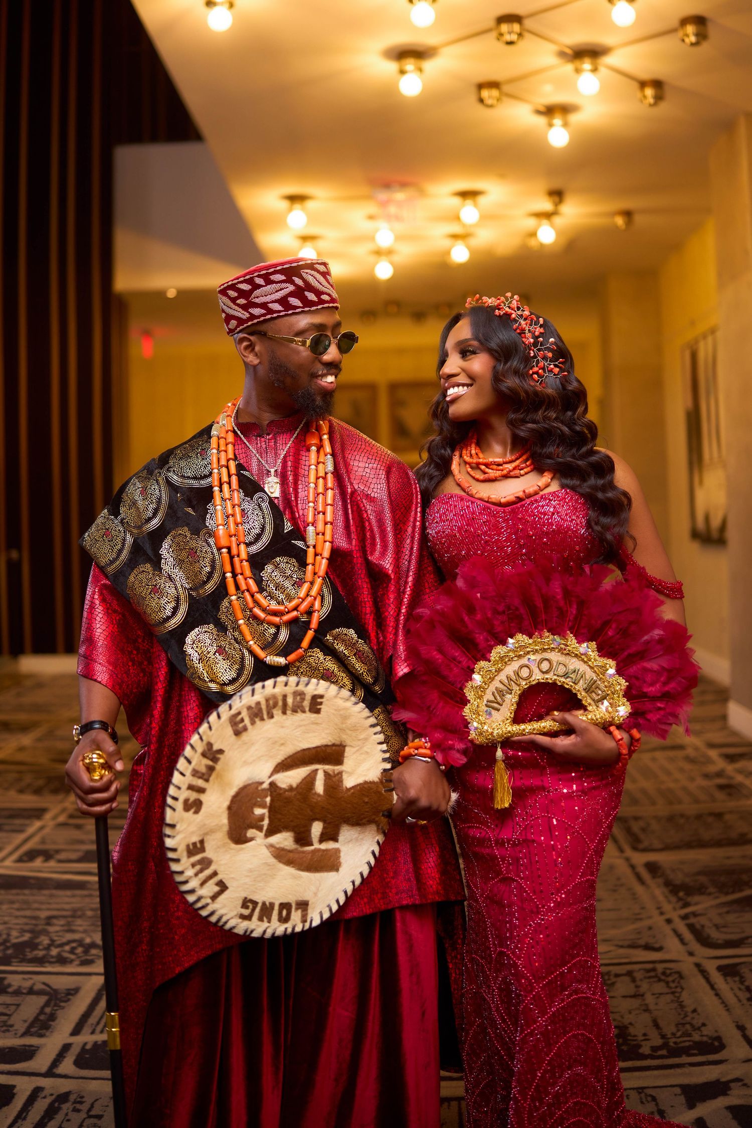 A couple in traditional Nigerian attire, with red and gold formal dress and beaded jewelry, poses together in a series.